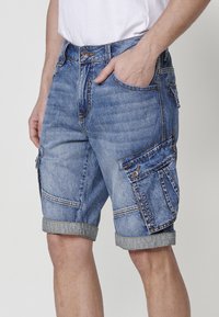 Lichtblauwe denim cargoshorts met omgeslagen rand, meerdere zakken en zichtbare stiksels. De stof lijkt gestructureerd en stevig.