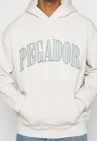 Felpa con cappuccio color beige chiaro con una grande stampa "PEGADOR" in verde-blu sul petto. Presenta una tasca frontale e polsini a costine.