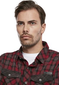 Rode en zwarte gecontroleerde flanellen overhemd met grijze accenten, knoopsluiting, twee borstzakken en een soepele textuur. Er onder een wit t-shirt.