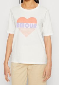 Kortärmad, krämfärgad t-shirt tillverkad av mjukt tyg som har en grafisk design av ett hjärta i korall och lavendel med ordet "AMOUR."