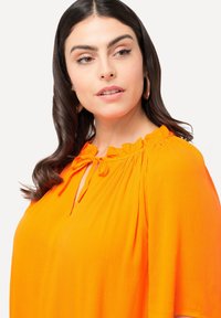 Blusa naranja brillante con un corte relajado, cuello con volantes y detalle de lazo. Tejido ligero con una textura sutil y mangas cortas.