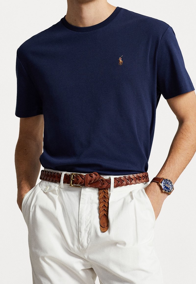 Navy cotton t-shirt with short sleeves, featuring a small embroidered logo. Paired with white trousers and a braided brown leather belt.