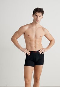 Fekete boxer alsónadrág piros gumis derékkal. Az anyag simának és rugalmasnak tűnik, szorosan illeszkedik a testhez, és jól látható első tasakkal rendelkezik.
