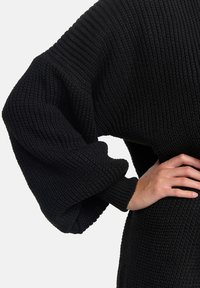 Svart stickad tröja med vida, ribbade ärmar och ett strukturerat mönster; tyget ser tjockt och varmt ut, med en avslappnad passform.