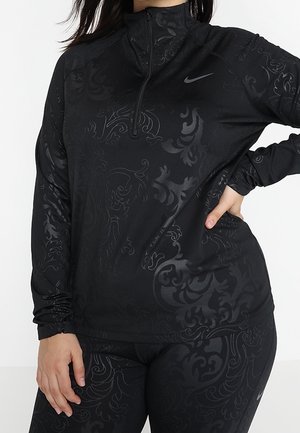 Kvinde iført sort Nike træningstøj med subtilt blomstermønster, langærmet trøje med lynlås og matchende leggings.