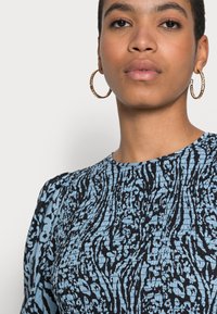 Top azul e preto com padrão e tecido texturizado. Apresenta um decote redondo e mangas curtas com volume. Brincos em forma de argola dourados complementam o visual.