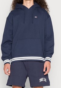 Navy hoodie i bomullsblandning, med en känguruficka, vita randiga muddar och en liten logotyp detalj på bröstet. Matchad med shorts i samma färg.