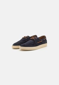 Navy blue suede shoes with beige rubber soles. Features brown laces and a decorative side tab. Rounded toe and simple design.