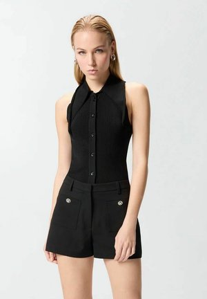 Giovane donna con capelli biondi pettinati all'indietro indossa un top nero senza maniche con bottoni e pantaloncini neri a vita alta con dettagli a tasche.