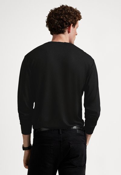 Fekete hosszú ujjú póló környakú szabással, puha anyagból, fekete farmerrel párosítva. Hátulról látható a lekerekített alsó szegély és a testhez simuló ujjak.