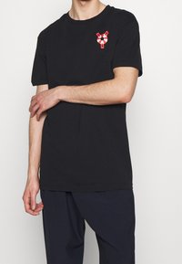 Černé bavlněné tričko s malým grafickým designem v červené a bílé barvě, který připomíná záchranný kruh na levé hrudi. Volný střih a krátké rukávy.