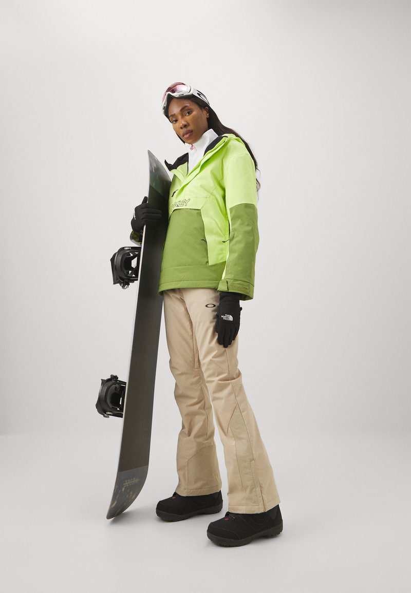 Grön och gul snöjacka, beige byxor, svarta handskar och stövlar, håller i en svart snowboard; runda goggles ovanpå mörkt hår.