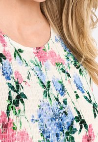 Top con motivo floreale e texture smock, caratterizzato da fiori rosa e blu su uno sfondo crema, scollatura tonda e maniche corte.