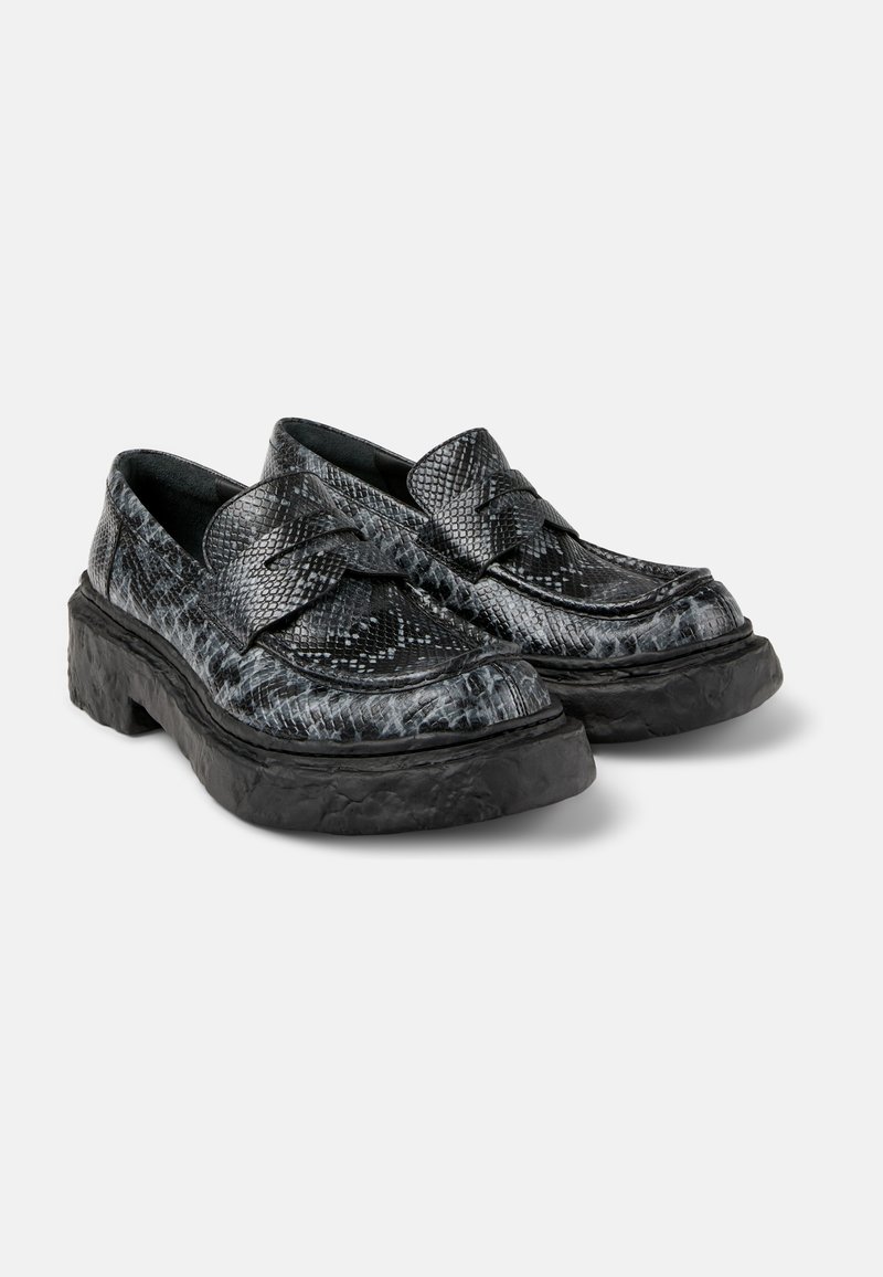 Black and grey textured shoes with a reptile pattern, featuring a thick black ridged sole and decorative bow. Designed for slip-on wear.