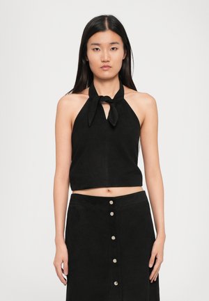 Jonge vrouw met lang, stijl haar in een zwarte haltertop geknoopt op de hals en een zwarte, knoopskirt, recht vooruit kijkend op een effen achtergrond.