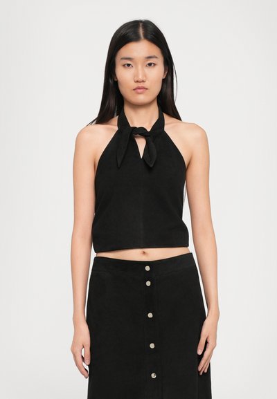 Jeune femme avec de longs cheveux raides portant un haut noir à lacets au cou et une jupe noire boutonnée, face à un fond uni.