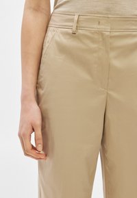 Αριστερό χέρι ακουμπισμένο δίπλα σε μπεζ παντελόνι ίσιας γραμμής με μπροστινές τσέπες και θηλιές για ζώνη, σε ουδέτερο φόντο.