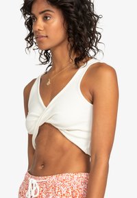 Biely krátky top s prekrúteným predným dizajnom. Hrebienková textúra, bez rukávov, spárovaný s patterned oranžovými šortkami s pásom na šnúrku.