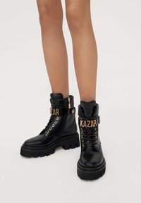 Botas de tobillo de cuero negro con suelas gruesas, detalles en dorado y un puño plegable. Presenta la marca "KAZAR" en una correa sobre los cordones.