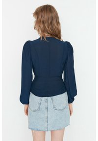 Navyblaues Langarmshirt aus strukturiertem Stoff, mit gepufften Schultern und taillierter Passform. Kombiniert mit einem hellen Denim-Minirock mit Gesäßtaschen.