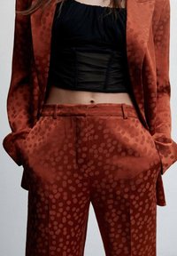 Costume de couleur rouille avec un motif à pois, comprenant une veste ajustée et un pantalon large. Tissu texturé avec poches latérales et passants de ceinture.