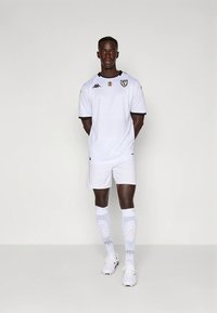 Bijeli atletski nogometni komplet koji uključuje kratku majicu s crnim detaljima i logotipom, odgovarajuće šortseve i bijele nogometne čarape do koljena s uzorkom.