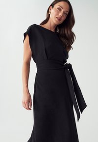 Musta mekko lyhyillä hihoilla, istuva bodice ja sidottu vyö, jossa on sileä pinta ja virtaava hame.