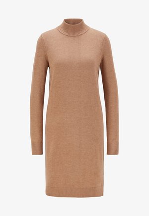 Kobieta ubrana w camelowy, długi sweterek i brązowe, zamszowe kozaki na obcasie do kolan stojąca na jednolitym tle.