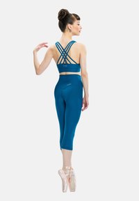 Ballet dancer in teal leggings and strappy crop top stands en pointe, facing sideways with hair in a bun against a plain white background.