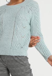 Jasnoniebieski sweter wykonany na drutach z teksturowanym, ażurowym wzorem. Posiada okrągły dekolt i ściągacz na dole, zestawiony z spodniami w kratę.