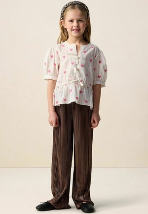 Blouse blanche avec motifs de cœurs roses et manches bouffantes, associée à un pantalon plissé marron. Le mannequin porte un serre-tête noir et des chaussures noires.