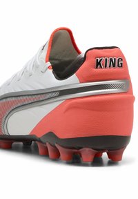 Clavado de fútbol en blanco y coral con parte superior texturizada, franja de acento plateada y suela negra con tacos rojos. "KING" impreso en el talón.