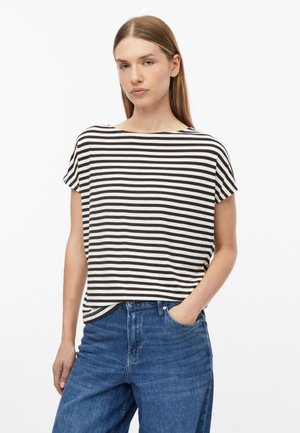 Jonge vrouw met recht lichtbruin haar, die een zwart-wit gestreept T-shirt met korte mouwen en blauwe jeans draagt, met één hand in de zak.