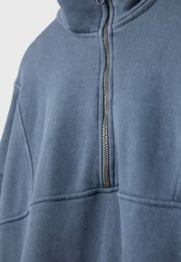 Gros plan d'une veste polaire bleue avec une fermeture éclair métallique verticale, montrant les détails des coutures et le tissu plié près du col.