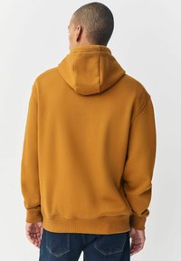 Sudadera color mostaza con un corte relajado, puños y dobladillo acanalados, y capucha. La tela parece suave con una textura lisa.