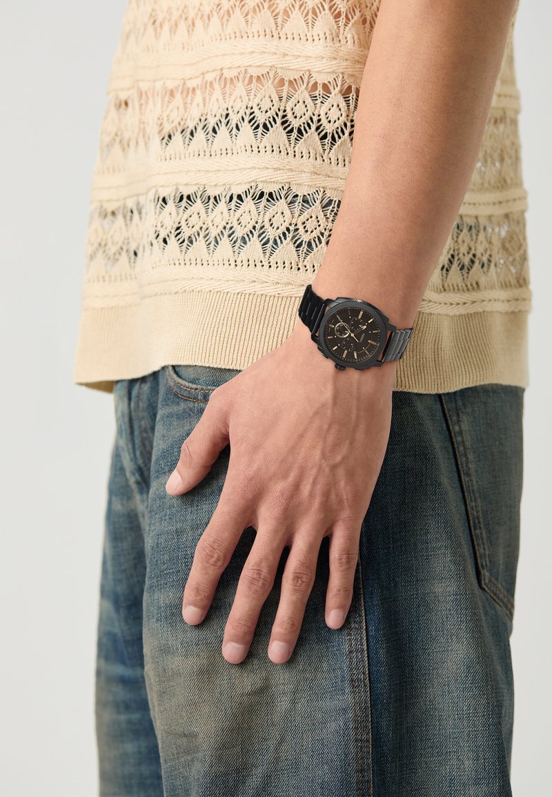 Montre analogique noire avec un boîtier texturé et un bracelet en caoutchouc, portée sur une main reposant sur un jean. Le haut en tricot présente un motif ajouré en beige.