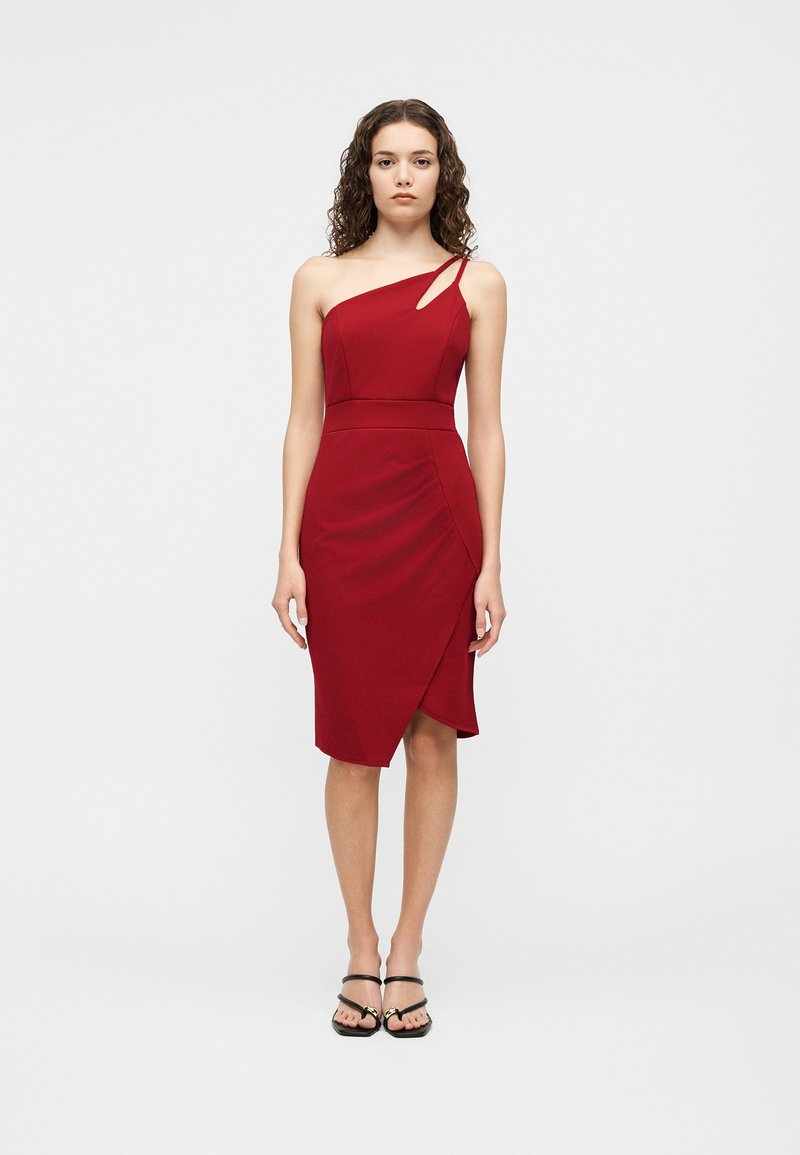 Vrouw met krullend haar die een nauwsluitende, knielange, asymmetrische rode jurk draagt met een klein uitsnijding en zwarte sandalen met bandjes.