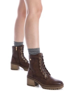 Botas de tobillo de cuero marrón con ojales dorados, tacón apilado y cremallera lateral. Combinadas con calcetines grises de canalé. Superficie texturizada en el calzado.