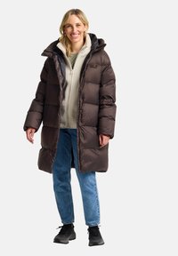 Bruine pufferjas met een capuchon, grote gewatteerde secties en drukknoopsluitingen, gelaagd over een beige fleece, gecombineerd met blauwe jeans en zwarte schoenen.