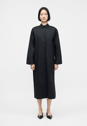 Woman standing wearing a long black buttoned dress with long sleeves and pointed black flats against a plain white background.