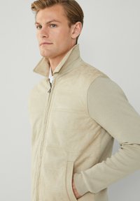 Bež atlet majica na zip s mekim, teksturiranim materijalom i prednjim džepovima. Ima visoki ovratnik i kombinaciju glatkih i rebrastih materijala.