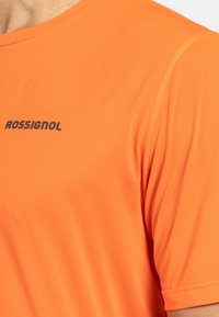 Oranžové sportovní tričko s kulatým výstřihem, krátkými rukávy a černým logem "ROSSIGNOL" na hrudi. Hladký, elastický materiál.