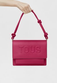 Fuchsia leather shoulder bag with embossed "TOUS" logo, knotted strap accents, and a flat design. Handheld with a smooth finish.