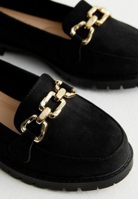 Black suede loafers with a gold chain accent, rounded toe, and a black rubber sole featuring textured tread for grip.