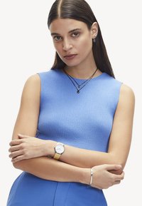 Blauw geribbeld tanktop, gecombineerd met gelaagde zwarte kettingen met hangers, zilveren horloge met gele band, zilveren armband.