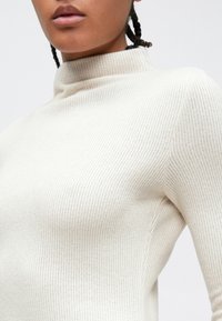 Pulover cu guler înalt, tricotat cu dungi, de culoare crem, cu croială strâmtă, oferind o textură fină și detalii subtile.