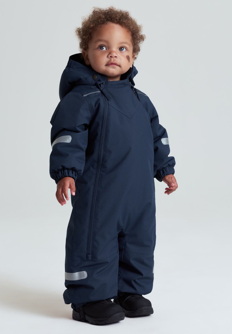 Småbarn med krøllet hår iført en mørkeblå isoleret flyverdragt med reflekterende striber og sorte støvler, stående mod en ensfarvet baggrund.