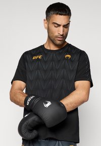 Μαύρο αθλητικό μπλουζάκι με υφασματικό μοτίβο, με το λογότυπο UFC. Φοριέται με μαύρα γάντια πυγμαχίας, που αναδεικνύουν τον κομψό σχεδιασμό και το κράτημα.