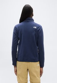 Σκούρο μπλε φλις πουλόβερ με ψηλό γιακά και λογότυπο στην πλάτη. Συνδυασμένο με ανοιχτό μπεζ παντελόνι, που παρουσιάζει τσέπες και ραφές λεπτομέρειες.