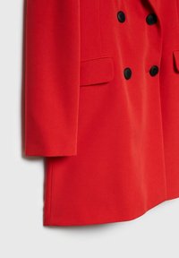 Manteau rouge croisé avec boutons noirs et poches à rabat, présenté de la moitié de la manche à l'ourlet sur fond blanc.